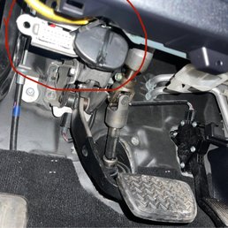 The photo depicts the driver side footwell of a vehicle, showing the accelerator pedal, brake pedal, and various mechanical and electronic components, including wiring and connectors.