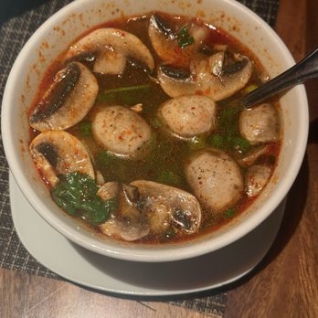 Sawasdee Danvers - Tom Yum - A bowl of Thai Tom Yum soup containing mushrooms, meatballs, and chopped green herbs in a spicy broth, served on a wooden table. - Thai Near Me - Danvers, Massachusetts