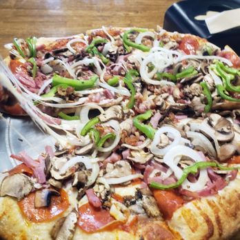 Pizza Factory - A large pizza topped with sliced mushrooms, onions, green peppers, ham, and pepperoni. The crust appears golden and slightly thick. The pizza is placed on a metal tray on a wooden table, with a black napkin dispenser visible in the background. - Pizza Near Me - Payson, Arizona