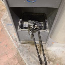 What is this??? - A gray ATM or vending machine with an open lower compartment holding various tools, keys, and a flashlight. The machine is positioned on a concrete floor with visible dirt and stains, and part of a brick surface is seen nearby. The top of the machine features a circular logo with a blue and white design.
