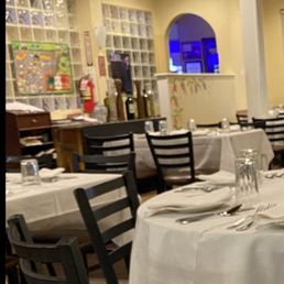 The restaurant interior features multiple tables with white tablecloths, each set with silverware, glasses, and napkins. Black chairs surround the tables, and the room has a decorative ceiling with patterned tiles. In the background, there is a counter area with bottles and a colorful wall display, and a small window with a blue-lit view beyond. The lighting is warm and ambient, creating a formal dining atmosphere.
