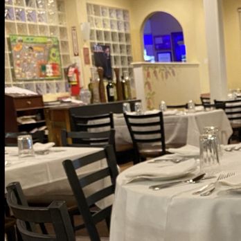 Pasquale Rigoletto Restaurant - The restaurant interior features multiple tables with white tablecloths, each set with silverware, glasses, and napkins. Black chairs surround the tables, and the room has a decorative ceiling with patterned tiles. In the background, there is a counter area with bottles and a colorful wall display, and a small window with a blue-lit view beyond. The lighting is warm and ambient, creating a formal dining atmosphere. - Italian Near Me - Bronx, New York