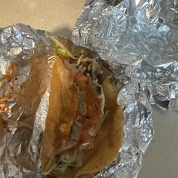 A taco with fried fish, lettuce, tomato, and sauce, partially unwrapped from aluminum foil, on a gray countertop.