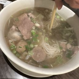 House Special Combo - A large bowl of Vietnamese pho containing beef slices, meatballs, rice noodles, and chopped green onions, with a hand stirring the broth.