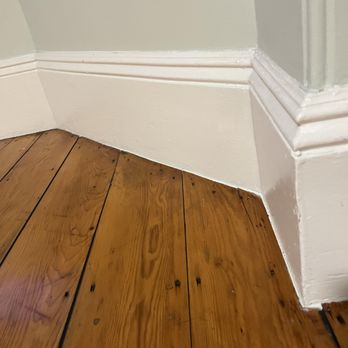 Advanced Flooring Solution - We worried that the black caulk might smear or stain our white baseboards, but they all look perfect. - A corner of a room features white painted baseboards with detailed molding, meeting at a right angle. The wooden floor consists of wide planks with visible grain, knots, and a warm finish. The baseboards are clean and well-painted, with some minor imperfections. The image emphasizes the transition between the wall and floor, showcasing the craftsmanship of the molding and the natural texture of the wood. - Flooring Near Me - Lowell, Massachusetts