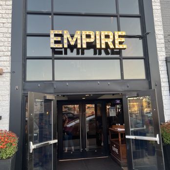 Empire - Front of restaurant - The image shows the front entrance of Empire restaurant, with a large glass window above the doorway displaying a lit sign spelling "EMPIRE" in marquee lights. The entrance has open double glass doors, and there are potted plants with flowers on either side of the doorway. The exterior features a white brick facade with black framing around the window and door. Reflections of parked cars and the street are visible in the glass. - Burgers Near Me - Naperville, Illinois