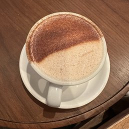 chai latte - A ceramic cup filled with chai latte featuring frothy milk and a layer of cinnamon powder on top is placed on a white saucer. The drink is on a wooden table with a smooth surface, and the image is taken from an overhead angle, showing the contrast between the frothy milk and the cinnamon dusting.