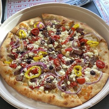 Bartlet Street - The best pizza ever. - A pizza with a thin crust is served on a white plate, topped with red onion slices, cherry tomatoes, yellow bell pepper slices, black olives, and crumbled cheese. The pizza is cut into slices and placed near a window with some papers and a menu in the background. - Pizza Near Me - Andover, Massachusetts
