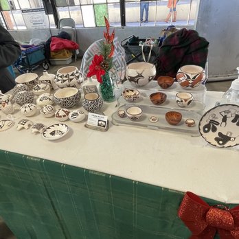 Rail Yards Market - A market stall features a variety of handcrafted pottery and ceramics, including bowls, plates, and decorative pieces with black and white geometric patterns and nature motifs such as reindeer and butterflies. The items are displayed on a white tablecloth, with a small green vase holding a red flower and pinecone decoration. A bottle of hand sanitizer is also visible on the table, and the background shows a market setting with other stalls and people. - Farmers Market Near Me - Albuquerque, New Mexico