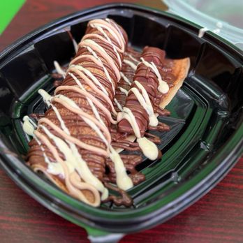Ranoush - Kinder Crepe - A chocolate crepe dessert is presented in a black plastic container, topped with a generous drizzle of white and milk chocolate. The crepe appears folded and is garnished with chocolate sauces, with a piece of pastry or cookie visible in the background. The container is placed on a wooden surface, and part of a clear plastic lid is visible in the background. - Waffles Near Me - Lexington, Kentucky