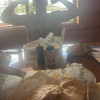 Subway - Tasty subway - A boy is seated at a wooden table, flexing his arms to show his muscles, with his face obscured by backlighting from a window with blinds. On the table, there are food items wrapped in paper, a salt or pepper shaker, a bottle of seasoning, and a bag of chips. The background shows a window with blinds, allowing sunlight to create a silhouette effect. - Sandwiches Near Me - Springerville, Arizona