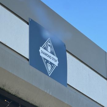 Diamond Stop - A rectangular blue sign featuring the Diamond Stop logo is mounted on the exterior wall of a building. The logo includes a diamond shape with a banner reading 'DIAMOND STOP' across it. The building's wall is light-colored with horizontal paneling, and the sky above is clear and blue. - Convenience Stores Near Me - Industry, Texas