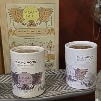 Kismet - Bath salts - Two cylindrical containers of botanical bath tea labeled 'Waxing Moon' and 'Full Moon' are displayed on a shelf, with a decorative 'Moon Bath' sign behind them. - Women's Clothing Near Me - Cincinnati, Ohio