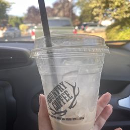 a hand holding a cup of coffee - A clear plastic cup containing iced coffee with some remaining liquid and ice cubes, held by a person's hand inside a car. The cup has a black straw and a logo that reads 'Country Coffee' with antler graphics. The background shows a parking lot with several cars and trees, with a partly cloudy sky.