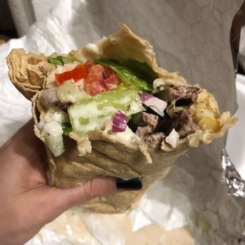 Barraco's - New York Steak Wrap - A person holds a partially eaten wrap containing lettuce, diced tomatoes, chopped onions, beef, and melted cheese. The wrap is wrapped in foil and paper, with some sauce visible at the bottom. The background includes a white textured surface and some paper towels, indicating a casual dining setting. - Pizza Near Me - Crestwood, Illinois