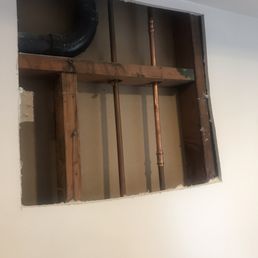 Before - A square section of an interior wall is cut out, exposing copper and black pipes along with wooden framing. The pipes are arranged vertically and horizontally, indicating plumbing work. The surrounding wall is painted white, and the opening provides a clear view of the building's internal structure and plumbing components.