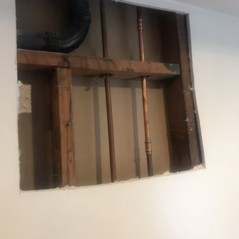 MRG Drywall - Before - A square section of an interior wall is cut out, exposing copper and black pipes along with wooden framing. The pipes are arranged vertically and horizontally, indicating plumbing work. The surrounding wall is painted white, and the opening provides a clear view of the building's internal structure and plumbing components. - Drywall Installation & Repair Near Me