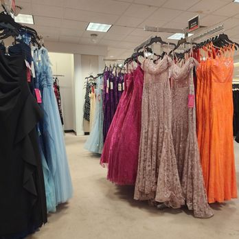 Dillard's - The image shows a retail clothing store with racks of formal dresses. The dresses are in various colors such as pink, beige, and orange, and are made of materials like lace and tulle. The store has a ceiling with fluorescent lighting and a beige carpeted floor. The dresses are displayed on black hangers, and the store appears organized with additional clothing racks visible in the background. - Department Stores Near Me - Plantation, Florida