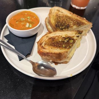 Scarlet Oak - Grilled cheese and tomato soup - A plate holds a grilled cheese sandwich cut in half, showing melted cheese and toasted bread with a golden-brown crust. A small bowl of tomato soup garnished with green onions is placed beside the sandwich. The plate is on a black table with a spoon, and drinks are visible in the background. The setting appears to be a restaurant or cafe. - New American Near Me - Washington, DC, District of Columbia