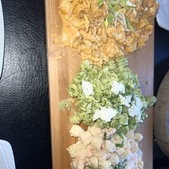 Dorsia - A wooden serving board displays three pasta dishes at Dorsia in Milwaukee, WI. The top dish appears to be a creamy pasta with mushrooms and green onions, the middle is green pasta with cheese and herbs, and the bottom is a pasta salad with vegetables and a creamy dressing. The dishes are presented side by side on the board, showcasing a variety of pasta styles and sauces. - Italian Near Me - Milwaukee, Wisconsin