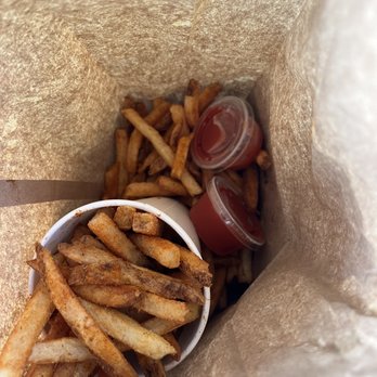 Five Guys - "Large" Cajun Fries - French fries with seasoning are placed inside a paper bag, accompanied by two small plastic containers of ketchup and a cup of fries. The fries are golden-brown and appear crispy. The image is taken from above, showing the fries and condiments inside the bag, which is lined with brown paper. - Burgers Near Me - Kingston, New York
