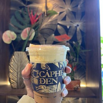 Caphe Den - Egg Cream Coffee - A hand is holding a plastic cup of coffee with a decorative sleeve that reads 'Caphe Den' in a cafe. The background features a patterned wallpaper with large leaf designs, a wall-mounted lamp, and potted plants, creating a cozy atmosphere. - Coffee & Tea Near Me - Flushing, New York