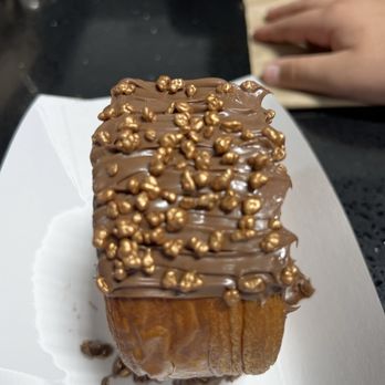 Baker 305 - A rectangular chocolate dessert with a smooth, glossy chocolate coating and gold-colored sprinkles on top. The dessert is on a white plate, with a black surface underneath and a hand holding a paper bag in the background. The image is focused on the dessert, highlighting its texture and decoration. - Bakeries Near Me - Miami, Florida