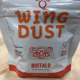 Kosmos Wing Dust - A 5-ounce bag of Kosmos Q Wing Dust Buffalo wing seasoning is shown, with branding and a buffalo illustration on the front. The packaging is primarily white with orange and red accents, and it is placed on a wooden surface. The bag promotes wing seasoning and includes phrases like 'Cook 'em naked!' and 'Dust 'em up!'.