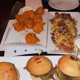 Boneless Wings - The image shows a table with plates of food including sliders with pickles, fried chicken pieces, and a pizza roll, with a person taking a photo.