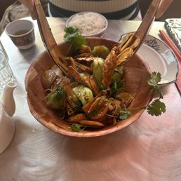 ML Dry Pot (build your own combo) - A person in a striped sweater is serving a stir-fried dish containing vegetables, seafood, and herbs from a large wooden bowl using tongs. The table has a bowl of white rice, a cup of tea, a teapot, and other dining utensils. The setting appears to be a restaurant with a casual dining atmosphere.