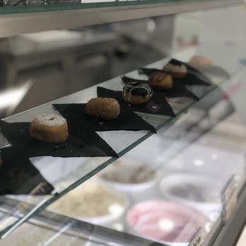 Y&E Creations - A row of mini donuts is displayed on black napkins inside a glass case at Y&E Creations in Middletown, NY. The donuts vary in flavor and appearance, with some topped with icing or dusted with powdered sugar. The background shows part of the shop's interior, including a glass shelf with decorative items and a menu or sign. The image emphasizes the freshly made donuts available at this sweet shop. - Desserts Near Me - Middletown, New York