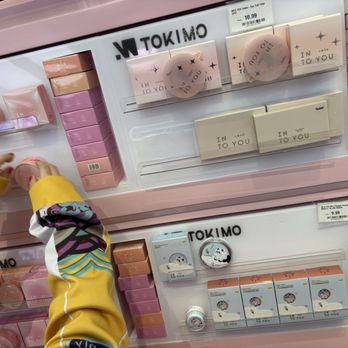 Tokimo - The image shows a retail display of skincare or cosmetic products at Tokimo in Flushing, NY. The display includes multiple boxes and containers in pastel pink, purple, and white colors, arranged on shelves. A person wearing a yellow and multicolored jacket is reaching for a product. Price tags are visible, indicating the products are for sale. The display is organized with branding and product packaging clearly visible. - Cosmetics & Beauty Supply Near Me - Flushing, New York