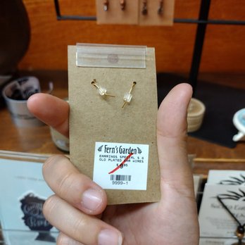 Fern's Garden - 50% off the marked price. eff 3/27/22 Sunday - A hand is holding a small cardboard package with earrings inside, featuring gold-colored posts and clear beads. The package has a label from Fern's Garden, a gift shop that sells handcrafted items. In the background, earrings are displayed on a wooden surface, and other shop items are partially visible. - Jewelry Near Me - Berkeley, California