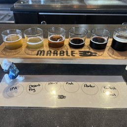 Flights - Six small glasses of beer, each with a different color and opacity, are placed on a wooden flight paddle labeled 'MARBLE' at Marble Brewery. A tasting note sheet with handwritten beer names is positioned below the paddle, indicating different beer styles such as IPA, Desert Fog, and Turkish Coffee. The background shows a bar counter surface.