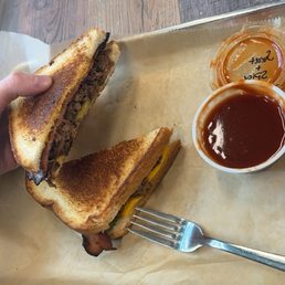 Brisket Stuffed Grilled Cheese. It had bacon and pickles on it and the sauce it came with was Amazing! - A grilled cheese sandwich with bacon and pickles is on a tray, accompanied by barbecue sauce and ketchup, with a fork nearby.