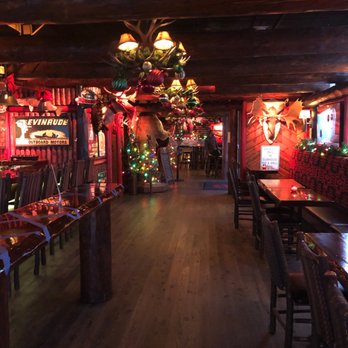 Camp Bar Third Ward - Atmosphere - The image shows the interior of a rustic-themed bar decorated for the holiday season. Christmas ornaments and string lights are hung from the ceiling and walls, including a mounted deer head with antlers. The decor includes a small Christmas tree with colorful lights and ornaments. The bar has wooden floors, tables, and chairs, creating a warm, cabin-like atmosphere. The lighting is dim, with some illuminated signs and decorations adding to the festive ambiance. - Bars Near Me - Milwaukee, Wisconsin