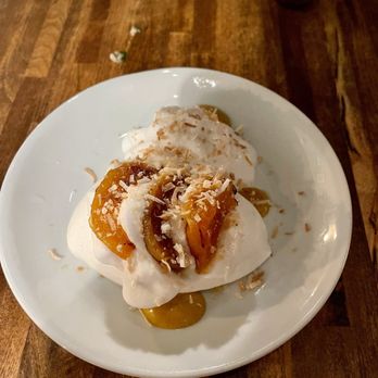 Thistle & Leek - Ginger pavlova - A ginger pavlova with whipped cream, caramelized fruit, and toasted coconut is served on a white plate, placed on a wooden table. - Gastropubs Near Me - Newton, Massachusetts