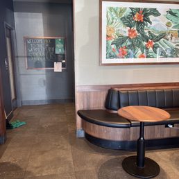 Inside a Starbucks, a black leather booth with a round wooden table is positioned against a wall with colorful botanical artwork. A chalkboard sign welcomes back customers.