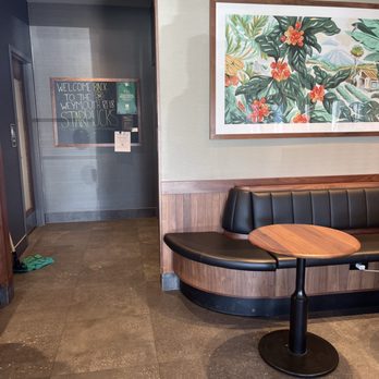 Starbucks - Inside a Starbucks, a black leather booth with a round wooden table is positioned against a wall with colorful botanical artwork. A chalkboard sign welcomes back customers. - Coffee & Tea Near Me - Weymouth, Massachusetts