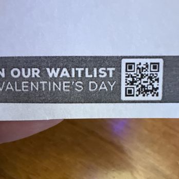 Texas Roadhouse - QR CODE - The image features a close-up of a printed paper, displaying a QR code and text related to joining a waitlist for Valentine's Day. The paper is held over a wooden surface, and part of the person's finger is visible at the bottom left corner. The text includes instructions to join the waitlist, with the QR code positioned to the right of the text. - Steakhouses Near Me - St. Augustine, Florida
