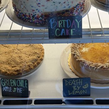Range Cafe - The image features three cakes inside a display case. The top cake is decorated with colorful sprinkles and a sign reading 'Birthday Cake.' The bottom left cake is labeled 'Green Chile Apple Pie,' and the bottom right cake is labeled 'Death by Lemon,' with a lemon glaze on top. The cakes are on white plates, and handwritten signs identify each dessert. - Breakfast & Brunch Near Me - Albuquerque, New Mexico
