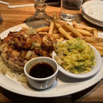 Cheddar's Scratch Kitchen - Key West Chicken & Shrimp - A restaurant plate contains grilled chicken and shrimp topped with sautéed vegetables, accompanied by a side of French fries and mashed potatoes with broccoli. The plate is set on a wooden table with glasses and plates in the background. - American Near Me - York, Pennsylvania