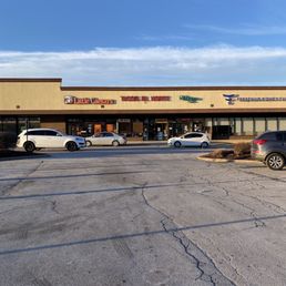 a parking lot with cars parked in it - A single-story strip mall features signs for Little Caesars, Tacos El Norte, and other businesses, with several parked cars in front. The parking lot has visible cracks and is mostly empty. The sky above is clear with some clouds, and the building has a beige facade with large glass windows and doors.