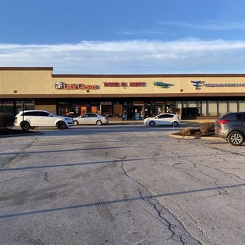Tacos El Norte - a parking lot with cars parked in it - A single-story strip mall features signs for Little Caesars, Tacos El Norte, and other businesses, with several parked cars in front. The parking lot has visible cracks and is mostly empty. The sky above is clear with some clouds, and the building has a beige facade with large glass windows and doors. - Mexican Near Me - McHenry, Illinois