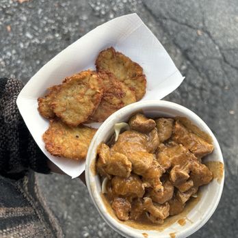 Reading Liederkranz - A hand holds a cup of beef stew with chunks of meat and gravy, along with fried plantains on a paper tray, outside on a paved surface. - Social Clubs Near Me - Reading, Pennsylvania