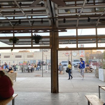 Gravity Bound Brewing - Looking outside - The image shows an outdoor dining area with several tables and people seated, viewed through an open garage-style door. Inside, part of the interior with seating and hanging plants is visible. The outdoor area has additional tables with patrons, and a building with a sushi restaurant sign is in the background. The scene suggests a casual dining environment with a mix of indoor and outdoor seating. - Brewpubs Near Me - Albuquerque, New Mexico