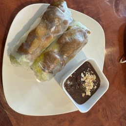 Spring Rolls - Two Vietnamese spring rolls, wrapped in translucent rice paper, are placed on a white square plate. The rolls contain visible ingredients such as lettuce and possibly other vegetables or proteins. A small square bowl of dark dipping sauce, garnished with crushed peanuts, accompanies the spring rolls. The plate is set on a wooden table, and the image captures a close-up view of the dish.