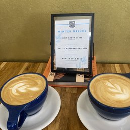 Toasted marshmallow and vanilla cinnamon - Two blue coffee cups with latte art sit on white saucers on a wooden table, with a menu sign displaying winter drinks behind them.