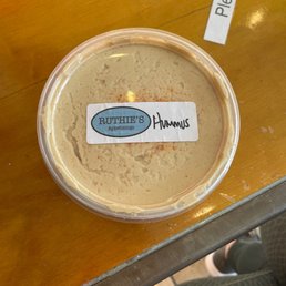 Hummus to go! Had to grab some for the week. - A clear plastic container filled with hummus, labeled with Ruthie's Appetizings, is positioned on a wooden table. The container has a white label with blue and black text, and the hummus appears smooth with a light beige color. A partially visible sign with the words 'Please do' is seen in the background, and part of a person's leg and shoe are visible at the bottom of the image.