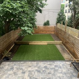 Final look! - A backyard enclosed by wooden fencing features artificial turf sections, a paved patio area, and a small bench, with trees and buildings in the background.
